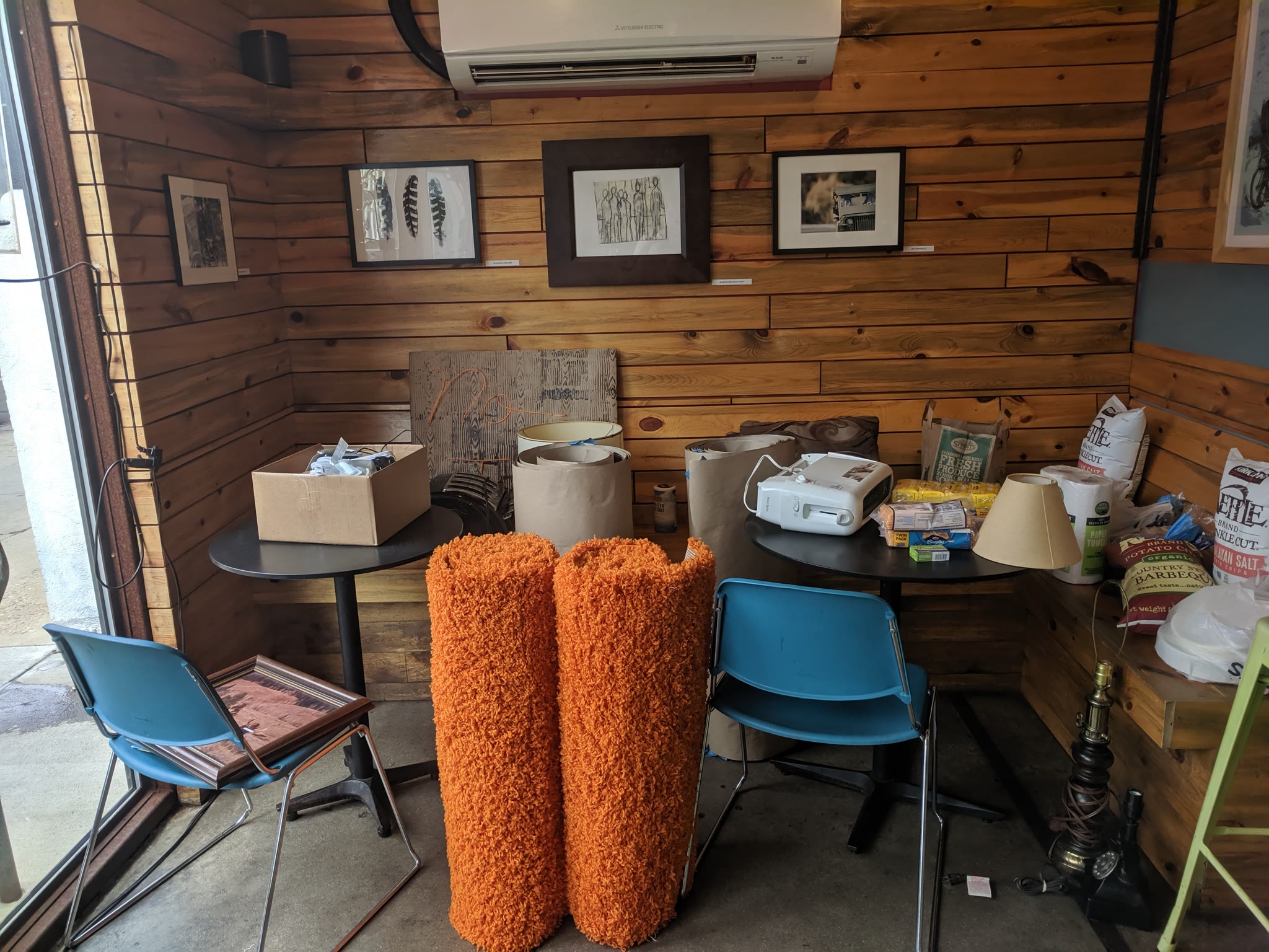 Upright rolls of orange shag rug, toasters, faux wood panel paper, and many other materials together in a cafe ready to be set up.
