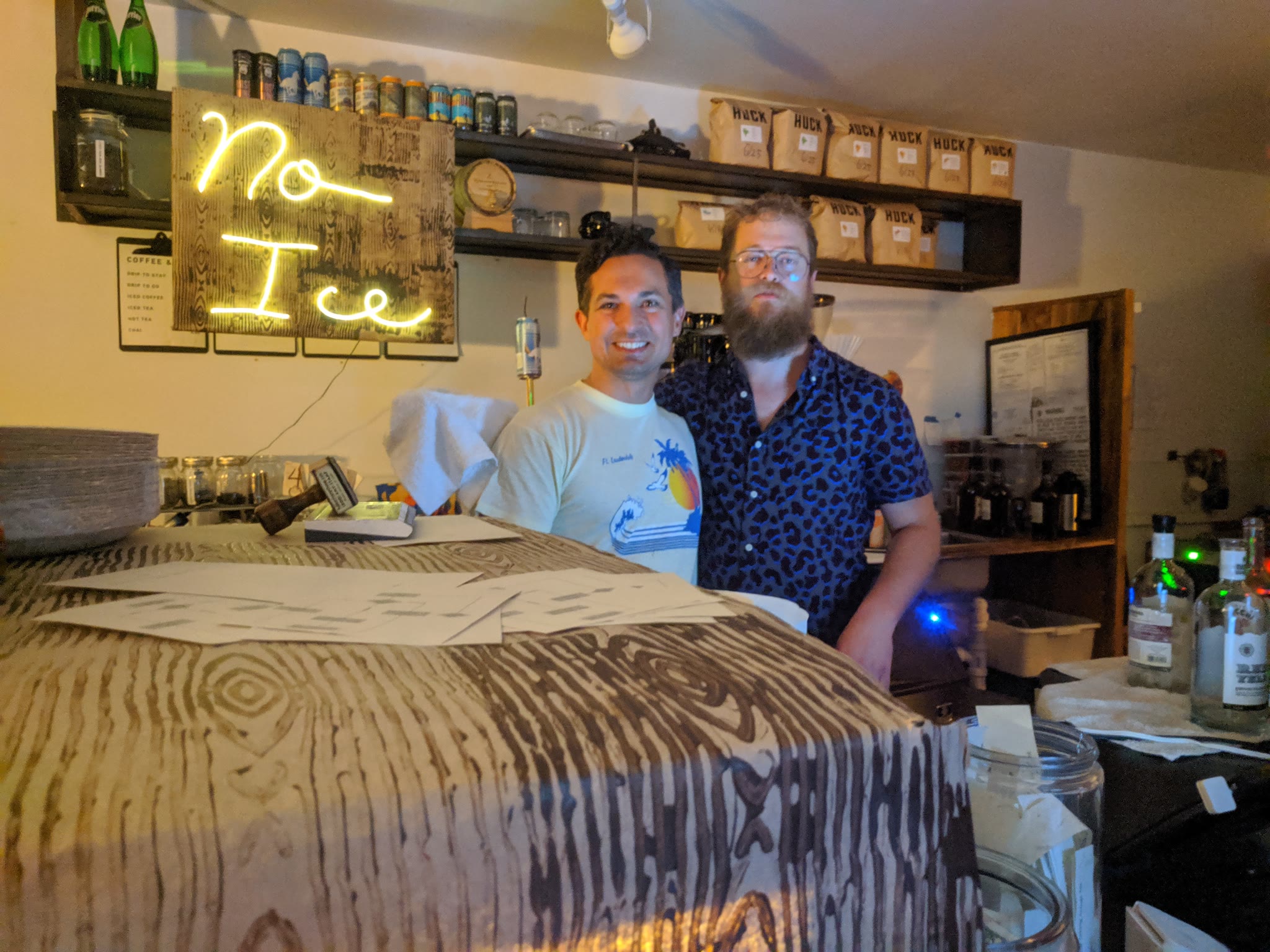 A picture of two men behind the bar at the No Ice event. The faux neon sign is lit up behind them. The person on the left, Wayne, is smiling. The person on the right, Neil, looks semi-serious.